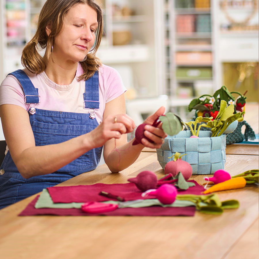 A woman wearing overalls making felt vegetables.